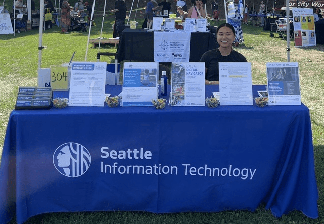Woman sitting behind a table with a blue tablecloth and five stands with paper in there. The tablecloth says Seattle Information Technology in white letters.