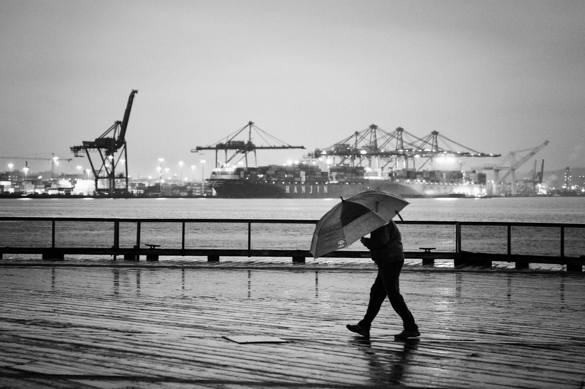 a man holding an umbrella along a shore with big ships in the background
