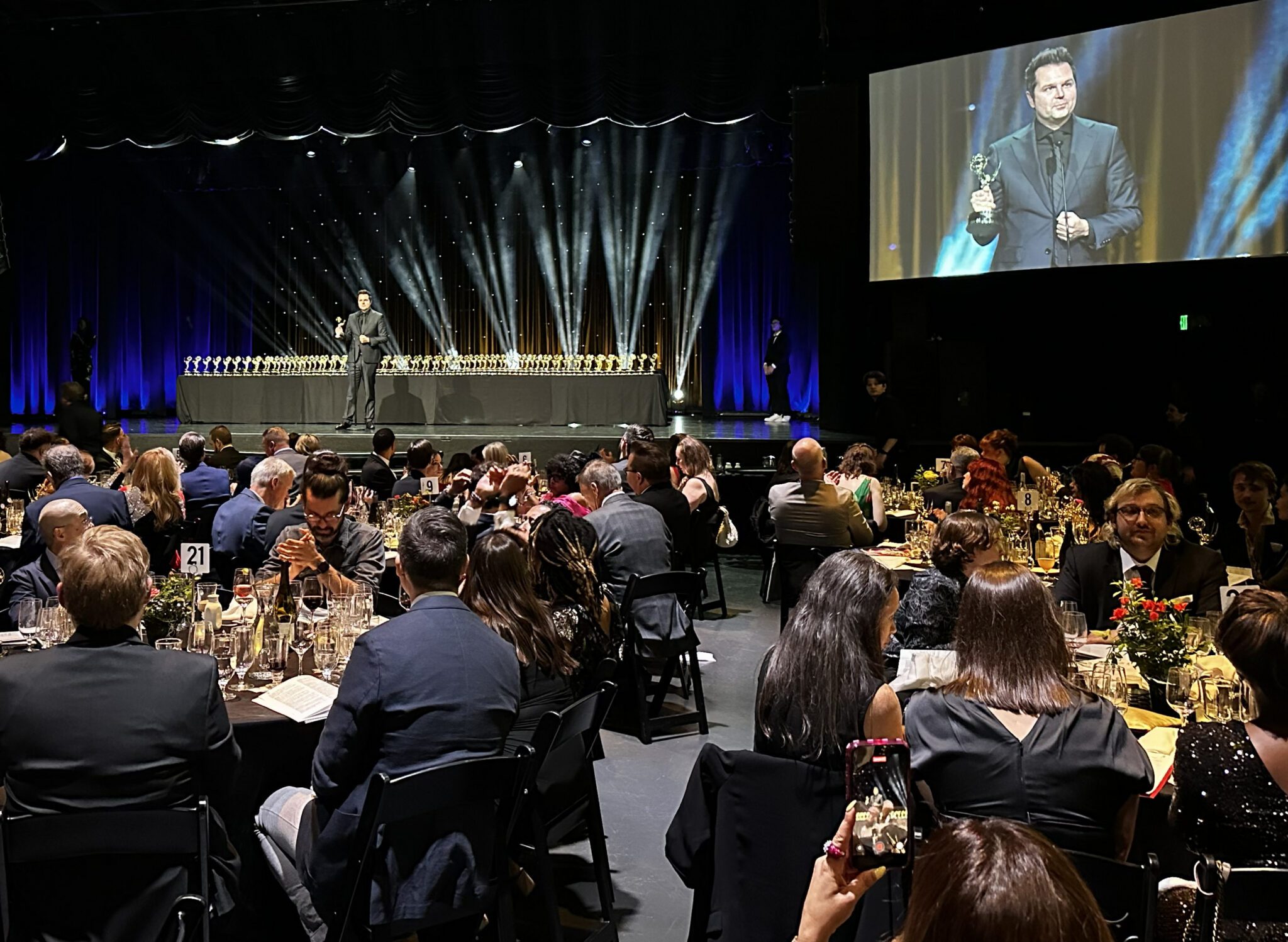 Stage of Regional Emmy awards with a man holding a golden Emmy award. People are sitting at tables watching the stage.