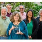 Eight people smiling and holding an Esri award outdoors, with green trees in the background.