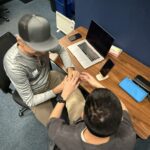 A man teaches a deafblind man how to use a laptop, guiding him through the keyboard with hands-on support.